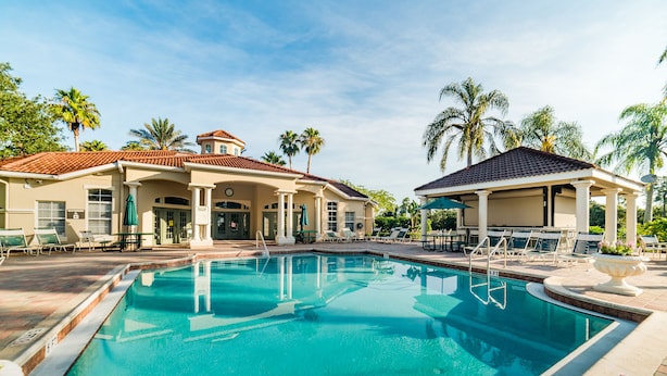 A swimming pool with lounge chairs and umbrellas, surrounded by palm trees and a building with a tiled roof.
