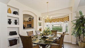A dining area with a glass-top table, wicker chairs, and a chandelier. There are shelves with decorative items and a large window with a view of the outdoors.