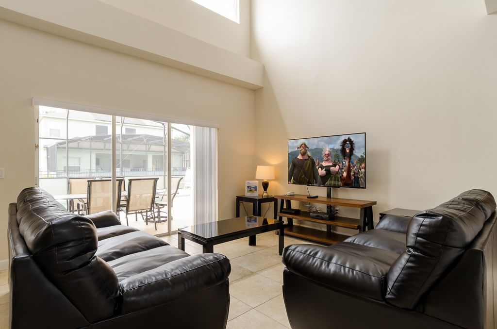 A living room with a black leather sofa set, a glass coffee table, a flat-screen TV mounted on the wall, and a sliding glass door leading to an outdoor patio area.