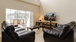 Un salon comprenant un ensemble de canapés en cuir noir, une table basse en verre, un téléviseur à écran plat fixé au mur et une porte-fenêtre coulissante donnant sur une terrasse extérieure.