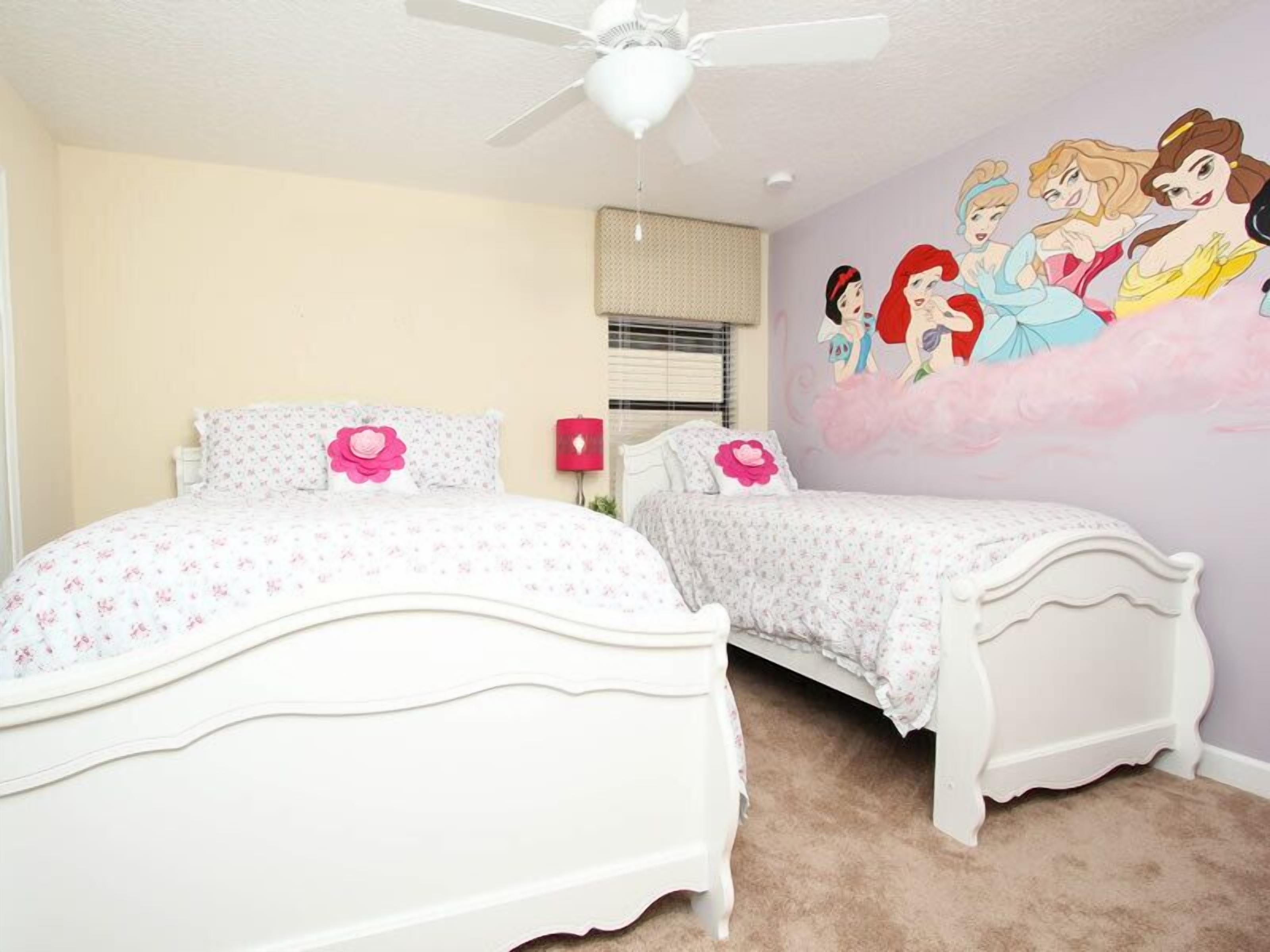 A bedroom with two white beds, a ceiling fan, a wall mural of Disney princesses, and a window with blinds.