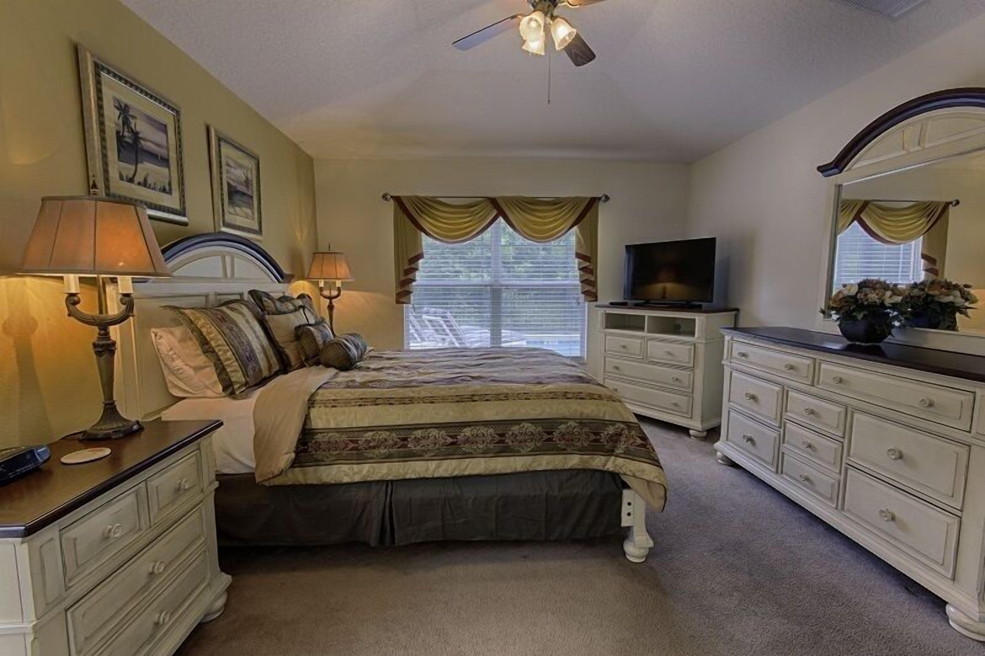 A bedroom with a bed, two bedside lamps, a television, a dresser, and a ceiling fan.