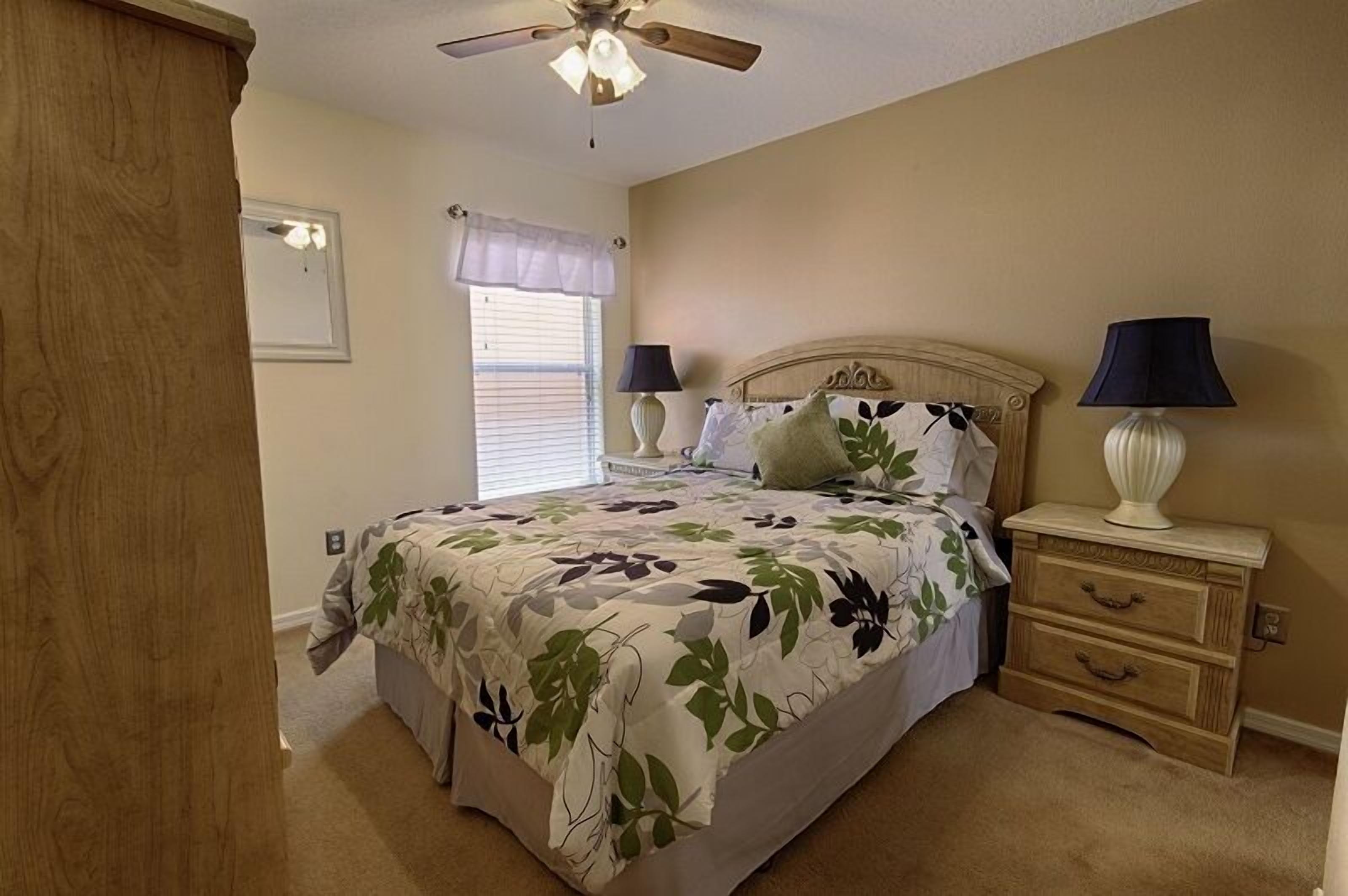 A bedroom with a bed, two bedside tables with lamps, a ceiling fan, a window with blinds, and a door.