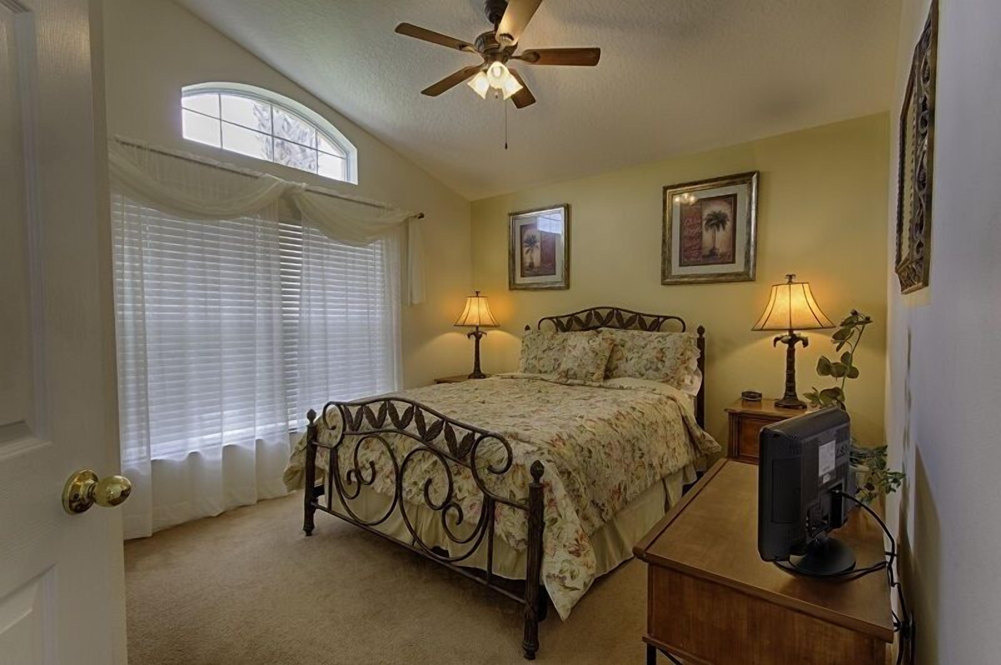 A bedroom with a bed, ceiling fan, two bedside lamps, a TV, and framed pictures on the wall.
