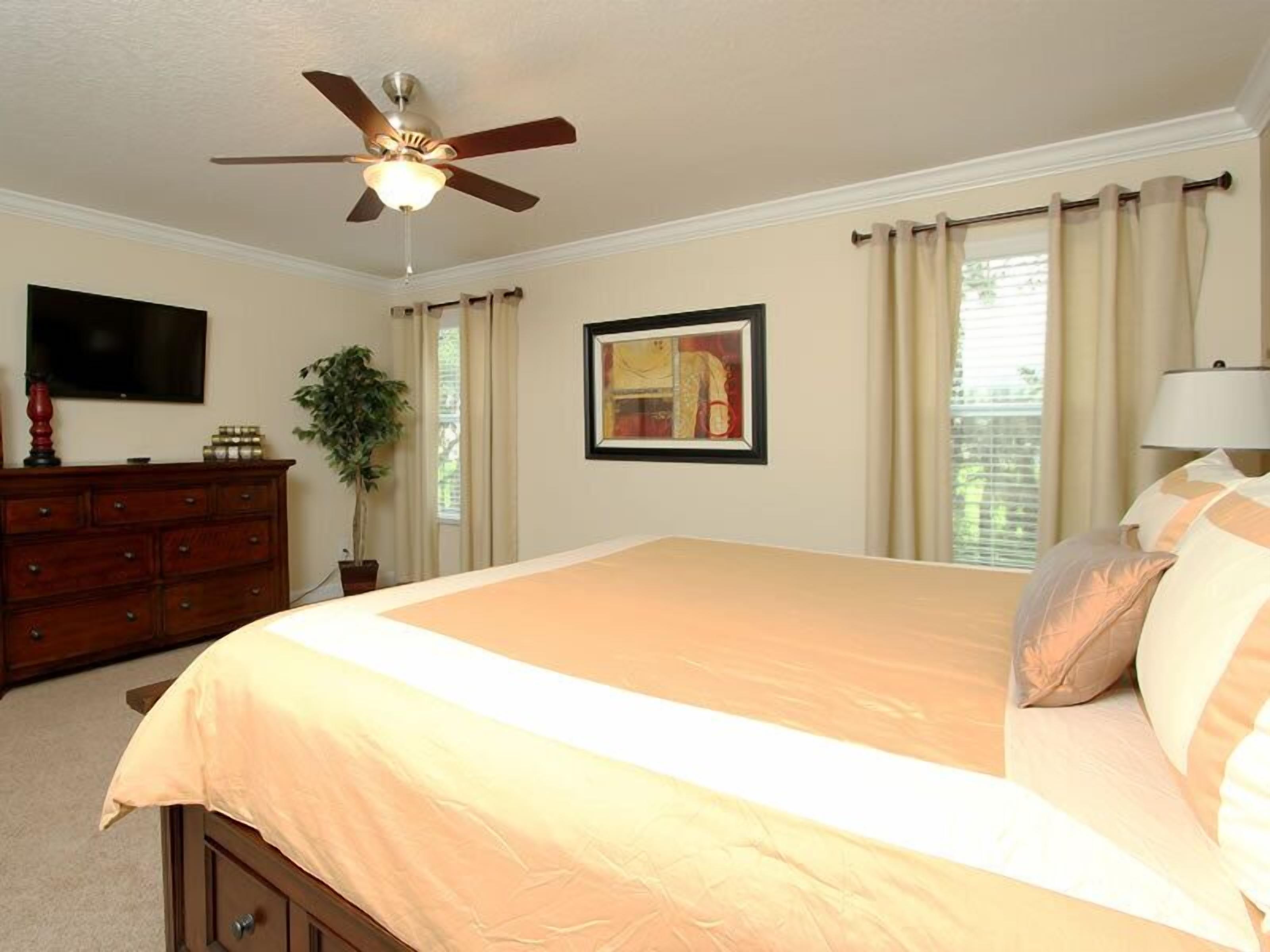 A bedroom with a bed, a ceiling fan, a dresser, a television, a window with curtains, and a framed picture on the wall.