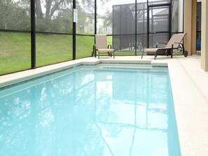 Ein rechteckiger, überdachter Swimmingpool mit klarem, blauem Wasser, umgeben von einer gefliesten Liegefläche. In der Nähe des Pools befinden sich zwei Liegestühle und ein kleiner Tisch.