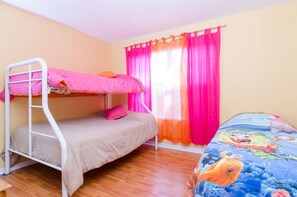Una habitación con una cama litera, una cama individual, un ventanal con cortinas rosadas y una colcha con un personaje de caricatura.