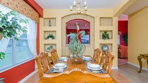 A dining room with a wooden table set for six, surrounded by chairs. There is a chandelier above the table, and the room has a red accent wall and a window with blinds.