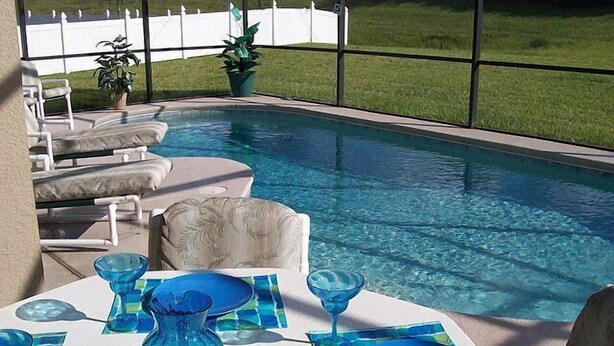 A poolside area with lounge chairs, a dining table set with blue glassware, and a view of a white fence and green lawn.