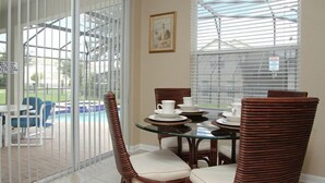 A dining area with wicker chairs and a glass table, a pool visible through a glass door, and a framed picture on the wall.