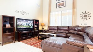A living room with a brown leather sofa, a television on a wooden stand, and a small table with a lamp. - Ovm604 - Emerald Island - 6 Bed 5.5 Baths Villa (Kissimmee)