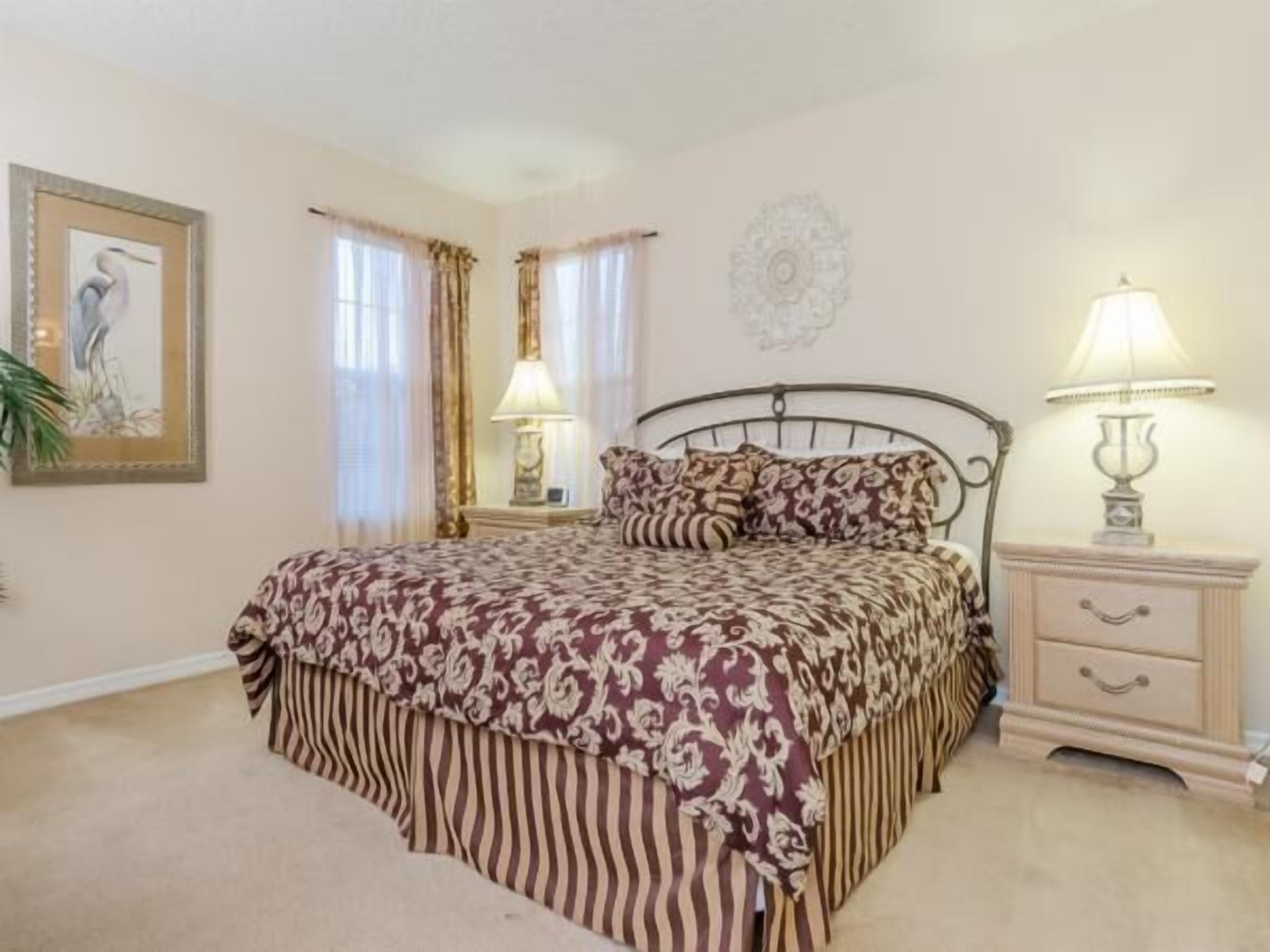 A bedroom with a bed, two bedside tables, a nightstand, a lamp, a framed picture, and a decorative wall piece.