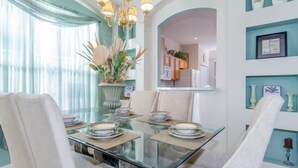A dining area with a glass table, white chairs, and a chandelier. There are decorative items on the walls and a view into the kitchen. - Ov535 - Emerald Island - 7 Bed 4 Baths Villa (Kissimmee)