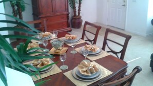 Una mesa de comedor dispuesta con platos, vasos y servilletas. Alrededor de la mesa hay sillas, un aparador de madera y plantas en macetas.