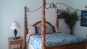 Una cama con dosel con cubrecama floral, una mesita de noche con lámpara y una planta en maceta en una habitación con pared lisa y un cuadro enmarcado.