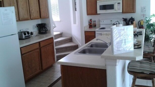 Una cocina con armarios de madera, encimera blanca, microondas, lavabo y una pequeña isla con una silla.