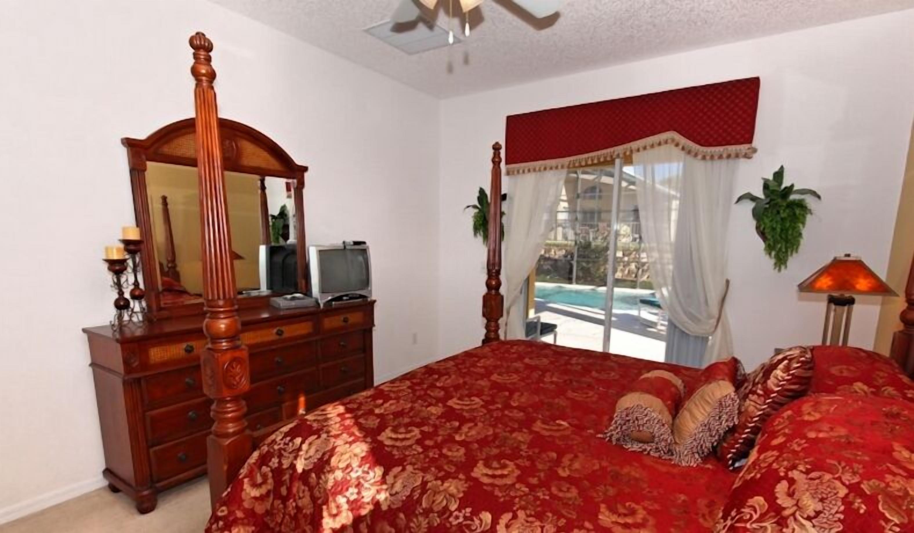 A bedroom with a four-poster bed, a wooden dresser, a television, and a view of a pool through a large window.