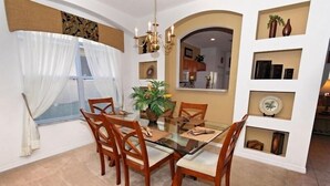 Un comedor con una mesa de vidrio y sillas de madera, una planta en maceta, y una cocina visible a través de un arco.