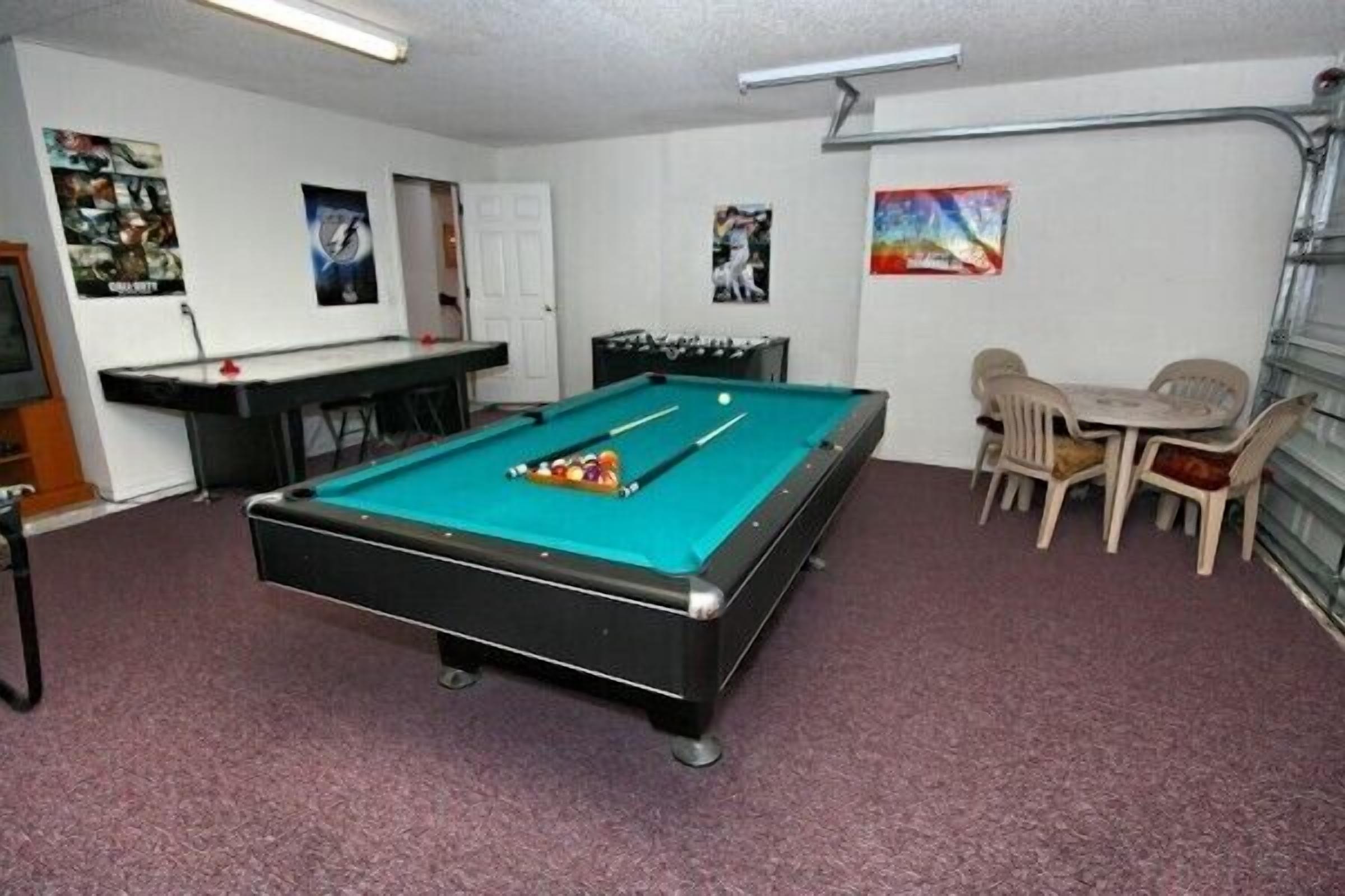 A pool table with a green felt surface, a foosball table, and a small seating area with plastic chairs.