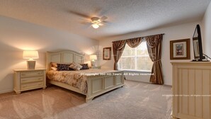 Ein Schlafzimmer mit einem Bett, zwei Nachttischen mit Lampen, einem Deckenventilator, einem Fenster mit Vorhängen und einem Fernseher auf einem Schrank.