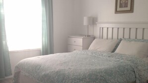 Quarto com cama, mesa de cabeceira, ventilador de teto e janela com cortinas.