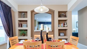 A dining area with a table set for four, surrounded by wooden chairs with a woven backrest. There is a chandelier above the table, and shelves with decorative items and books on either side. - Ov1138 - Emerald Island - 7 Bed 4.5 Baths Villa (Kissimmee)
