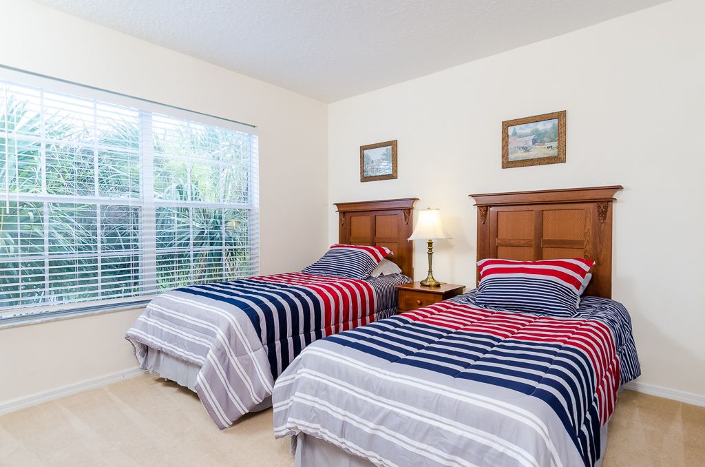 A bedroom with two beds, a window with blinds, and a nightstand with a lamp.