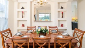 Een eetkamer met een grote houten tafel, zes stoelen, een kroonluchter en planken met decoratieve objecten.