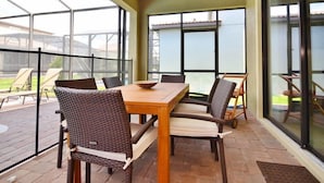 Une terrasse couverte avec une table en bois et des chaises en osier, de grandes fenêtres et une vue sur un immeuble.