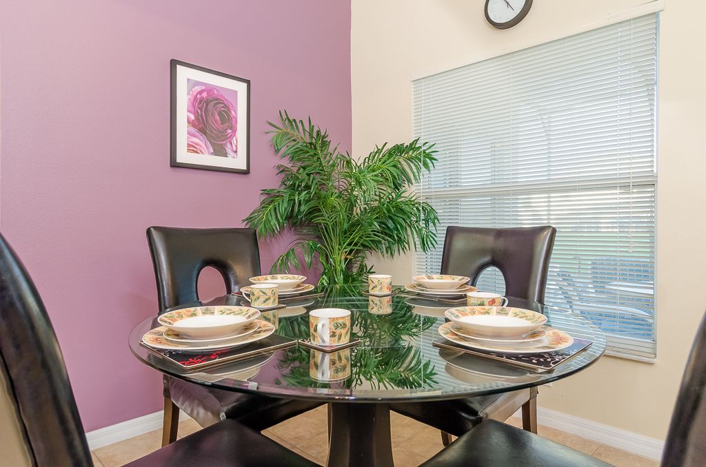 A dining area with a glass table set for four, surrounded by black chairs, a potted plant, and a framed picture on the wall.