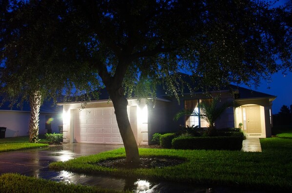 Une maison d’habitation de nuit, avec un garage, une allée et un arbre au premier plan.
