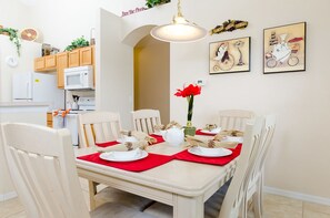 Un coin repas avec une table dressée pour quatre personnes, des chaises blanches, des sets de table rouges et un vase rempli de fleurs rouges.