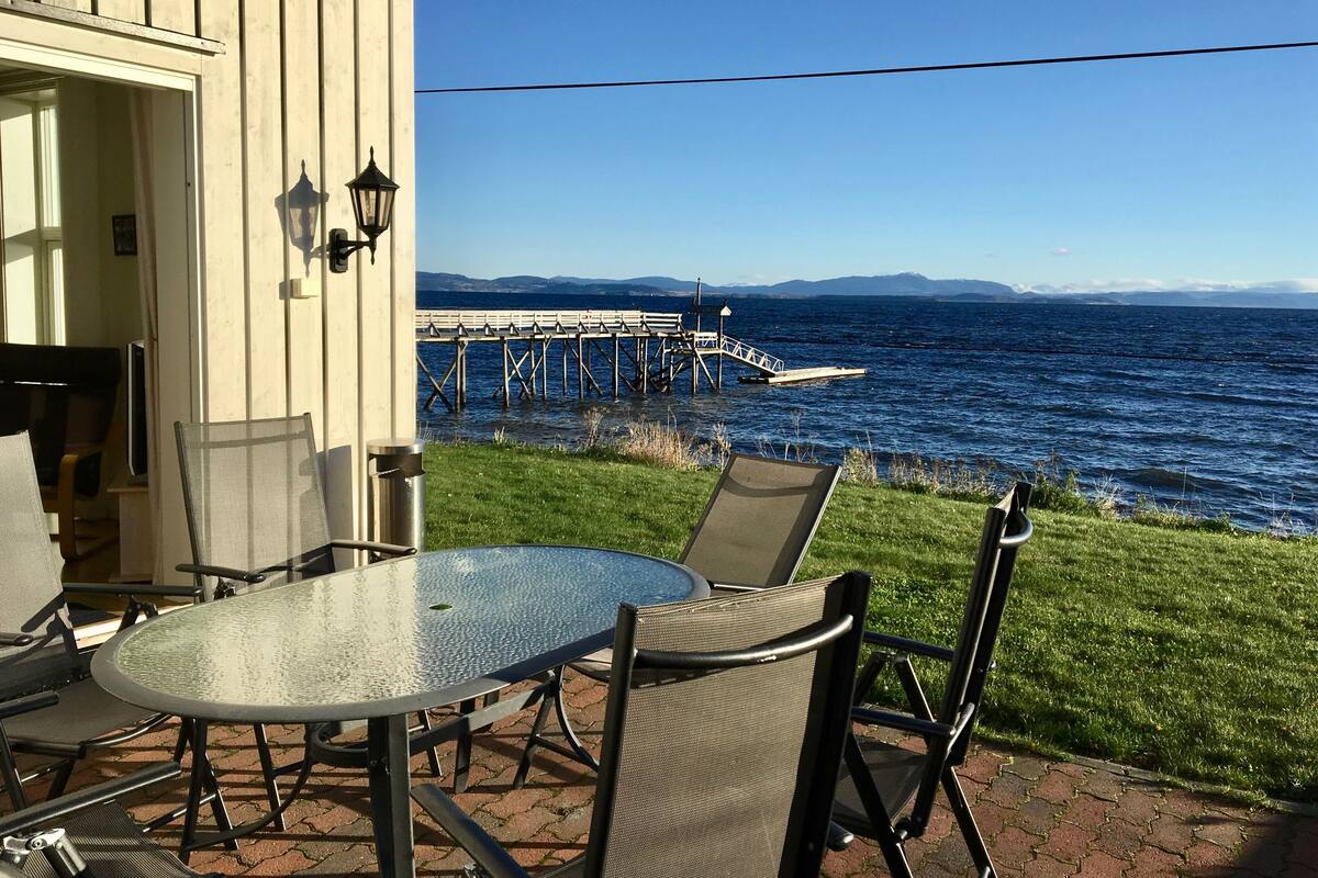 Un patio al aire libre con una mesa redonda de vidrio y sillas, una terraza de madera, y vista al océano con un muelle al fondo.