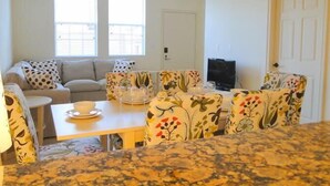 Un salon avec un canapé, une table basse et un téléviseur. Il y a deux fenêtres et un ventilateur de plafond.