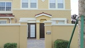 Un bâtiment à deux étages, doté d’une façade beige, d’une porte noire et d’un petit jardin agrémenté d’arbustes taillés.