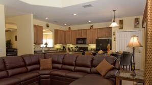 A modern living room with a sectional sofa, a dining area with a table and chairs, and a kitchen with wooden cabinets and appliances.