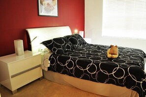 Un lit bien fait, recouvert d’une couvre-lit à motifs noir et blanc, une table de chevet blanche avec une lampe, et un tableau encadré accroché au mur.