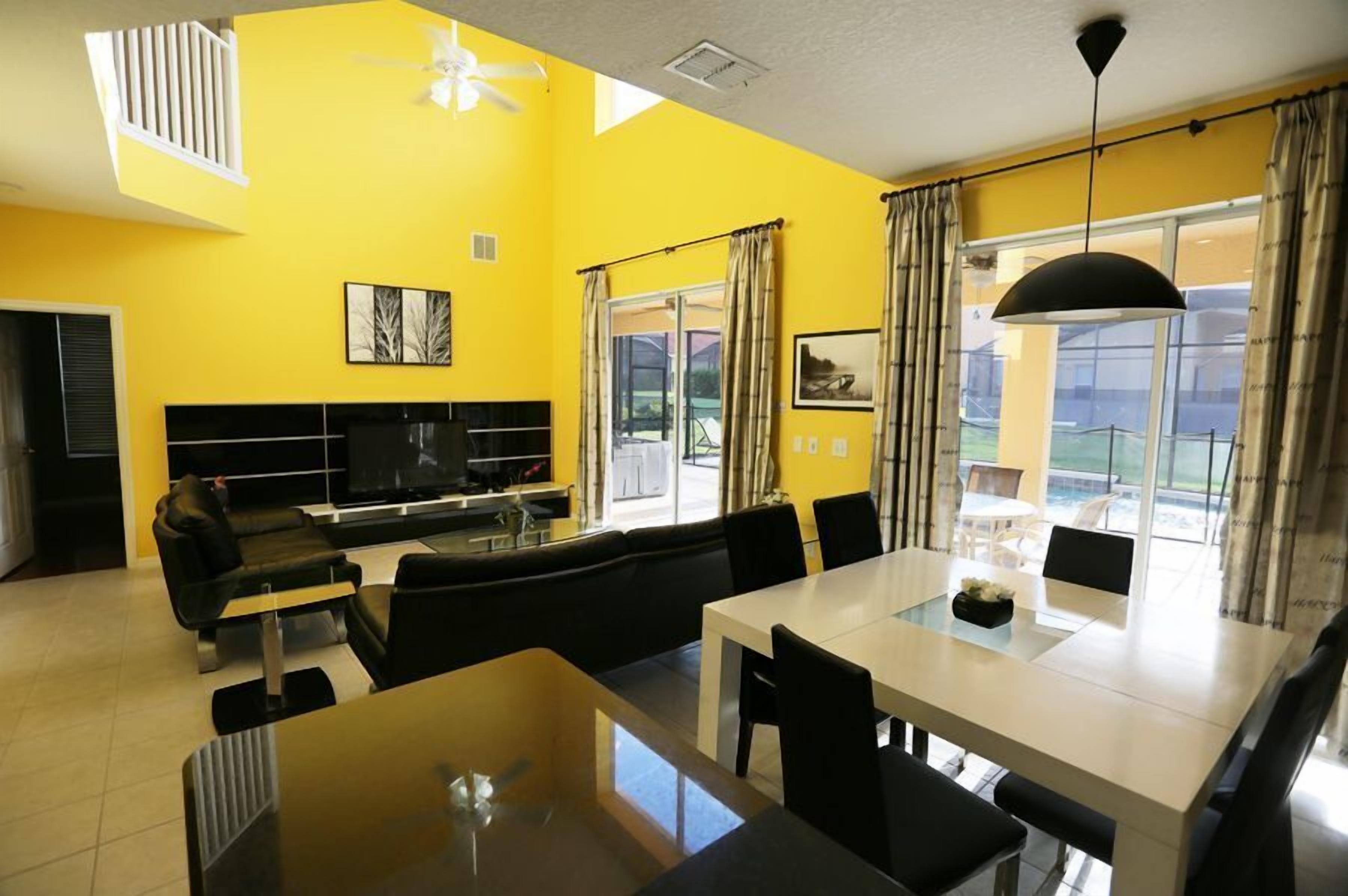 A modern living room with a yellow wall, a black sofa, a dining table, and a TV.