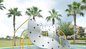 Un parque infantil con tobogán y estructuras para escalar, rodeado de palmeras y jardines.