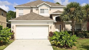 A two-story house with a white garage door, a small attached garage, and a well-maintained front yard with palm trees and shrubs.