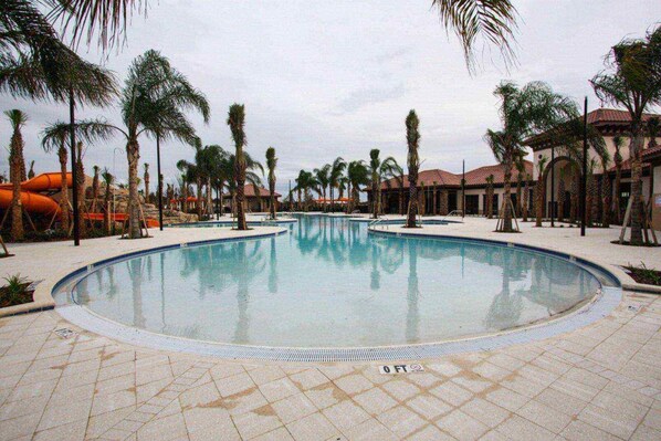 A large, clear swimming pool surrounded by palm trees and buildings with red-tiled roofs.