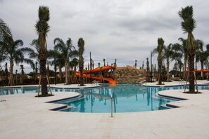 A swimming pool with a water slide, surrounded by palm trees and a stone wall.