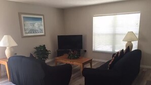 Un salon avec deux canapés noirs, une table basse en bois, un téléviseur à écran plat, une plante en pot et une fenêtre avec des stores.