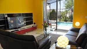 A modern living room with a glass coffee table, a black leather sofa, and a TV on a red stand. - Ov3990 - Emerald Island - 7 Bed 4.5 Baths Villa (Kissimmee)