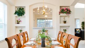 Un coin repas avec une table en bois dressée pour six personnes, entourée de chaises, avec un lustre au-dessus et des plantes en pot disposées sur des étagères.