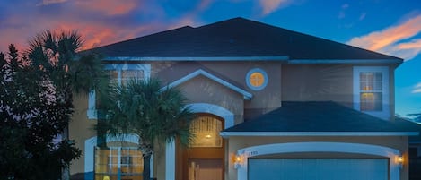 A two-story house with a garage, a circular window on the second floor, and a well-maintained front yard with palm trees.