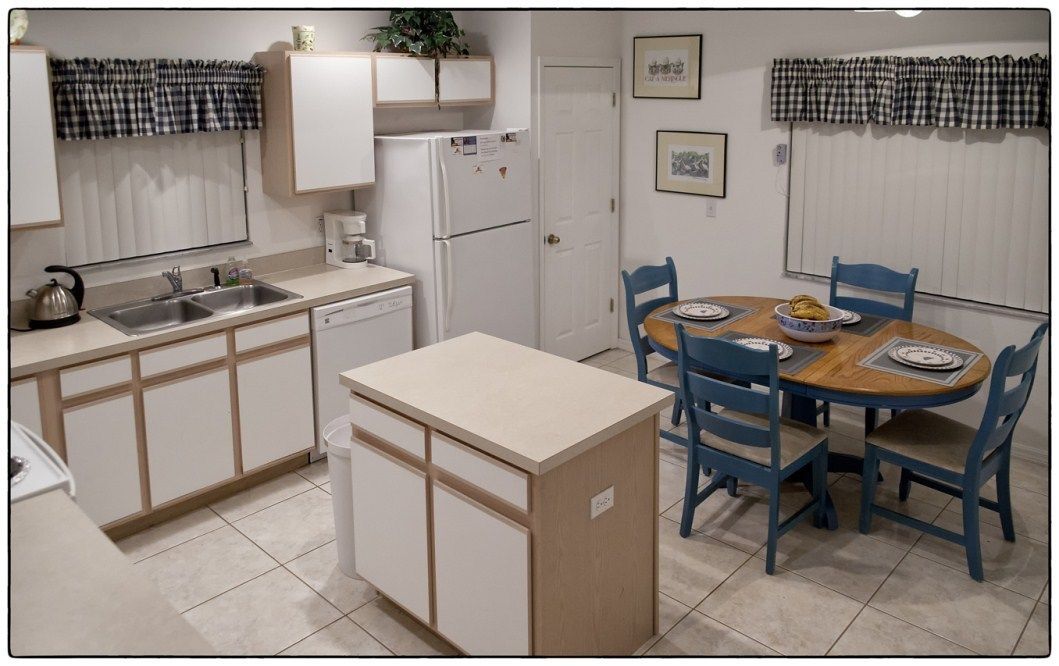 A compact kitchen with white cabinets, a sink, a coffee maker, and a refrigerator. There is a dining table with blue chairs and a small island with a sink.