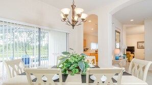 Un coin repas avec une table dressée pour quatre personnes, entouré de chaises blanches, d’un lustre et d’une grande fenêtre munie de stores.