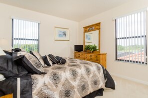 Quarto com cama, penteadeira, televisão e duas janelas com persianas.