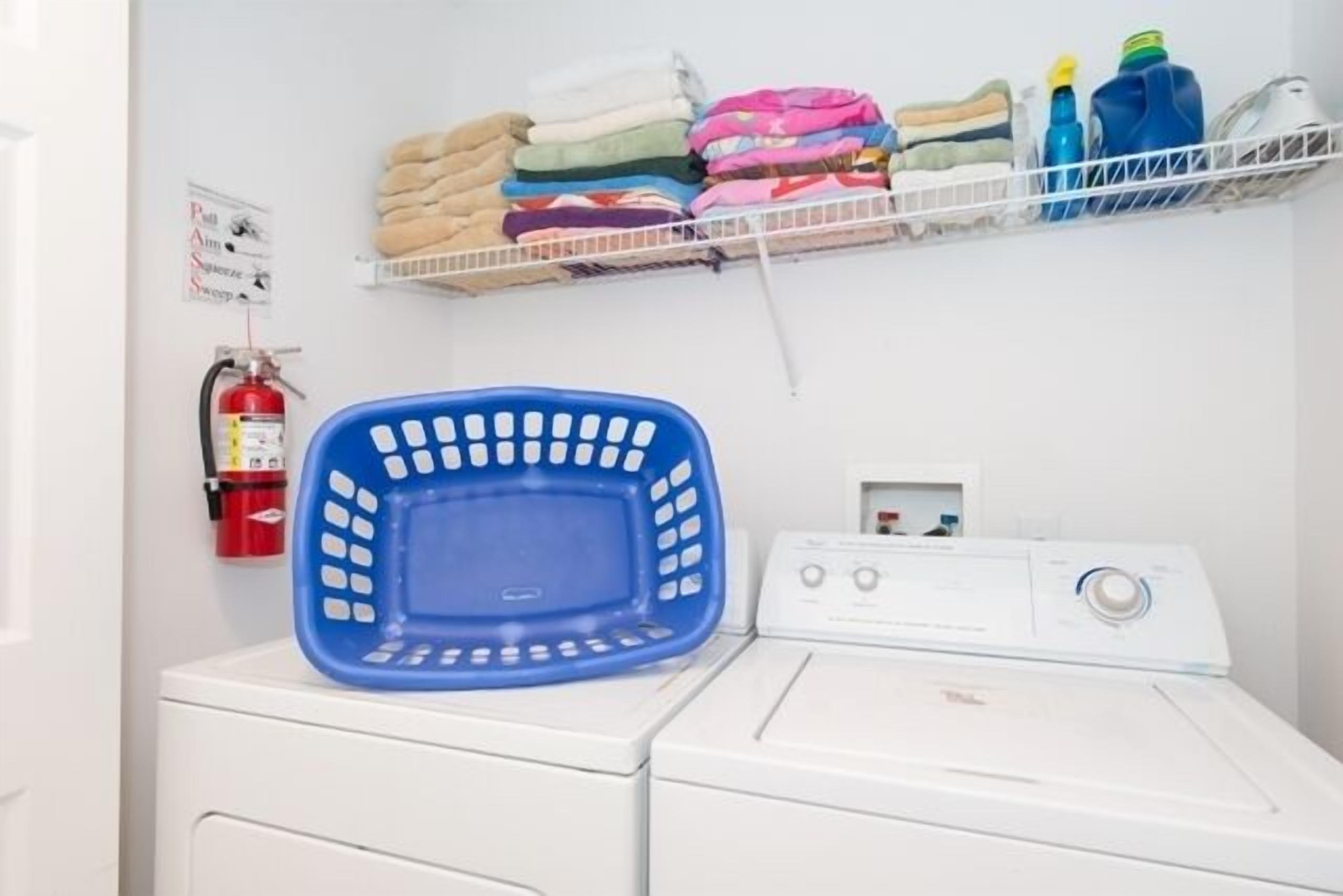 A laundry area with a washing machine, a blue laundry basket, and a shelf with folded clothes and cleaning supplies.